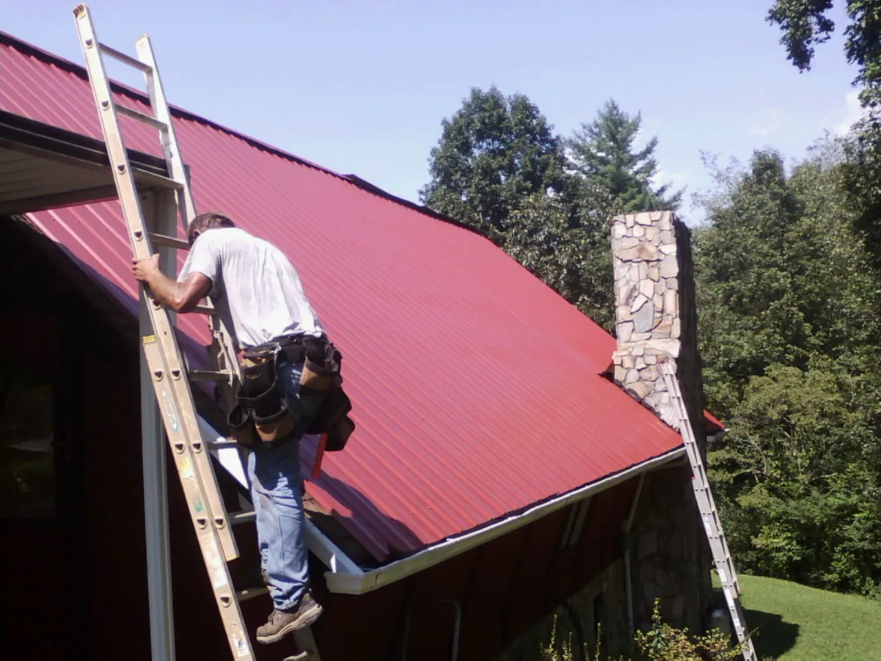 Roofer on a ladder beside a red metal roof and stone chimney — Soffit & Fascia Repair by Roofing Brothers in Mishicot